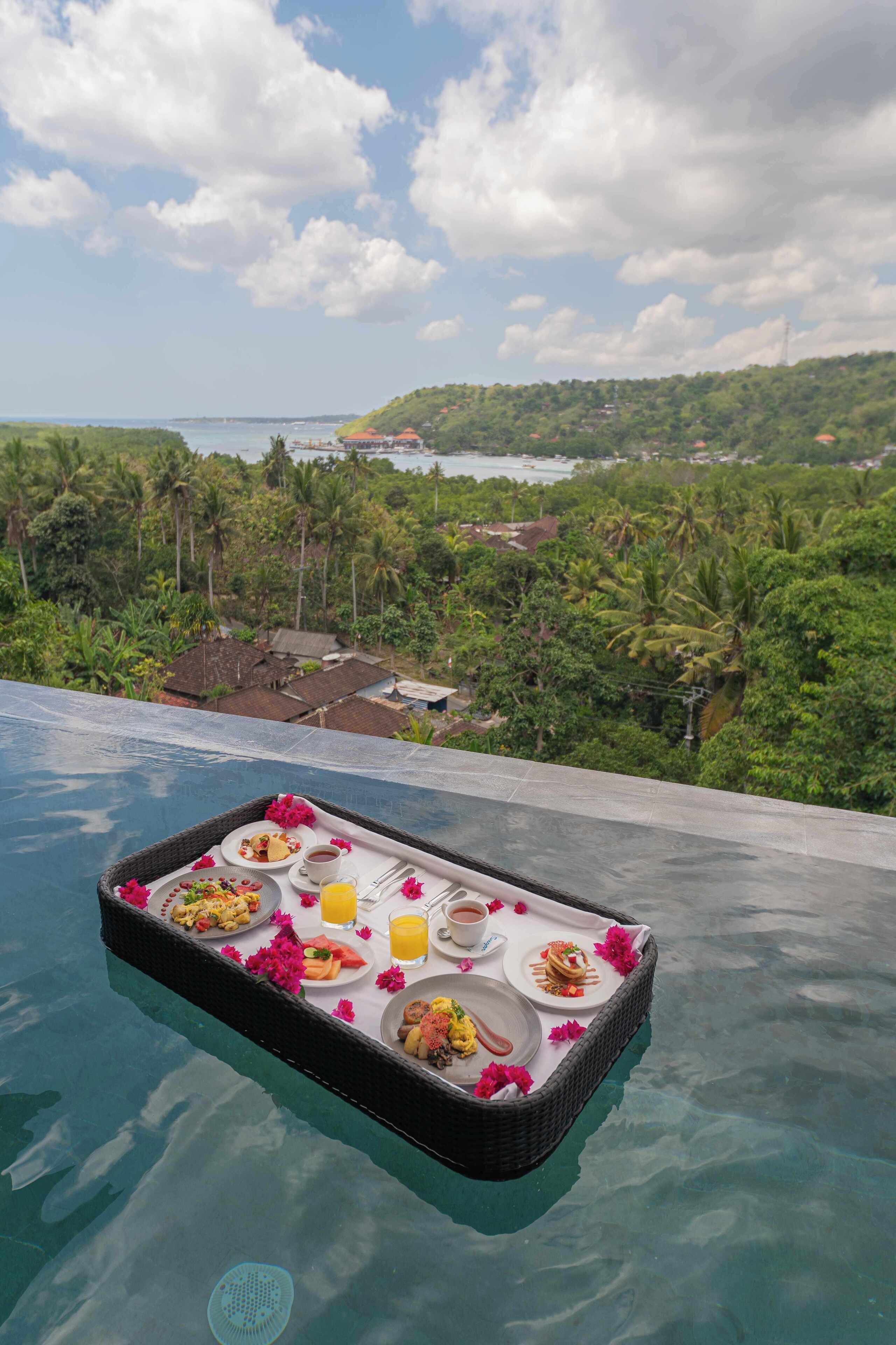A floating tray with breakfast items over a pool, surrounded by lush greenery and a view of a coastal area.