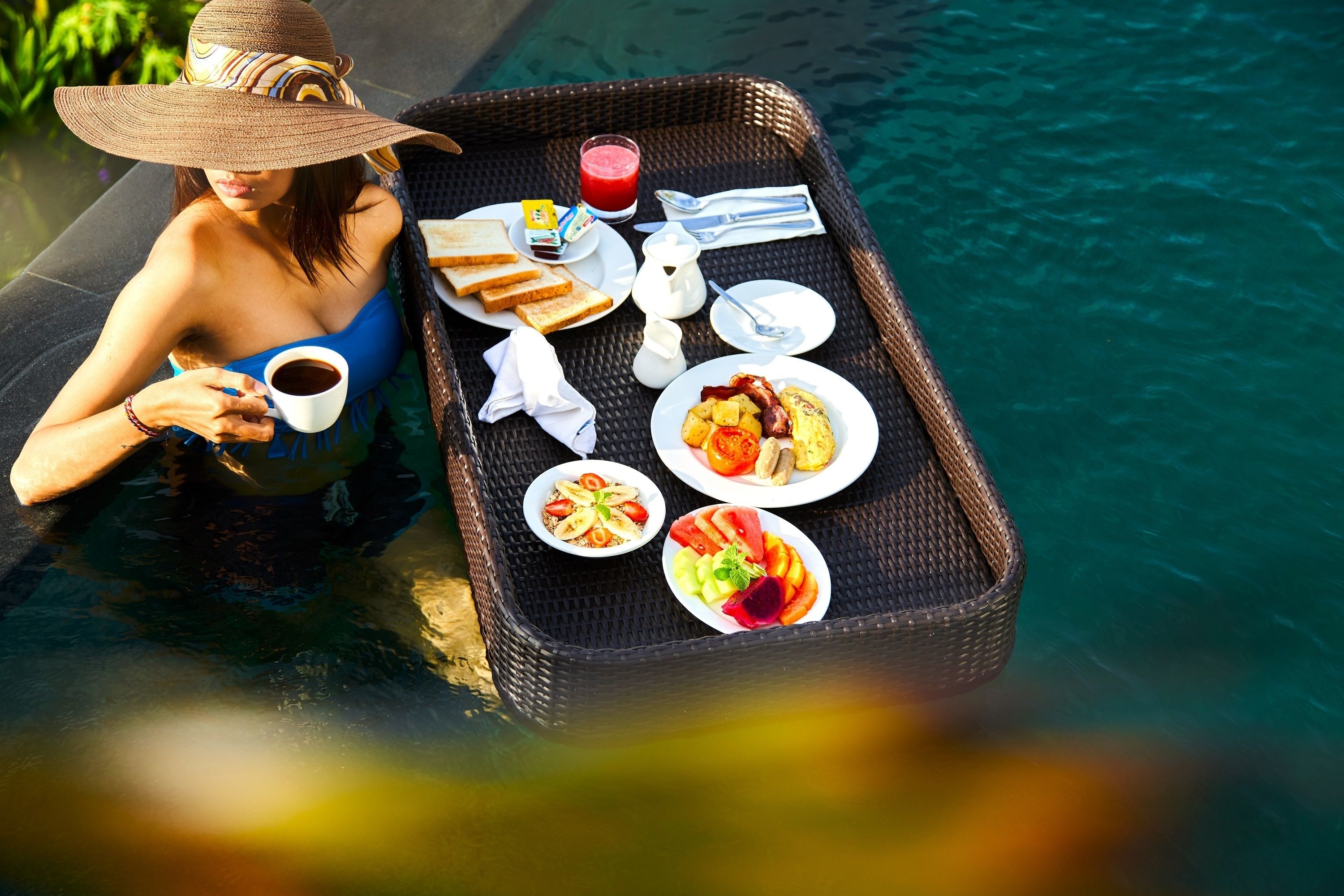 Bandeja de mimbre con alimentos para el desayuno, como tostadas, huevos y fruta, colocada junto a una piscina.