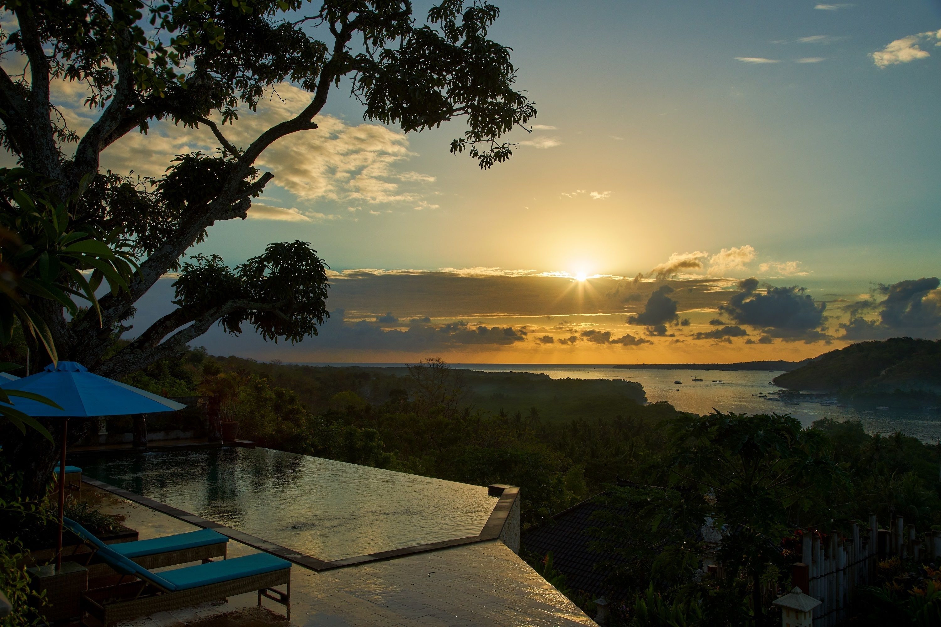 A infinity pool with a view of the sunset over a body of water, surrounded by lush greenery.