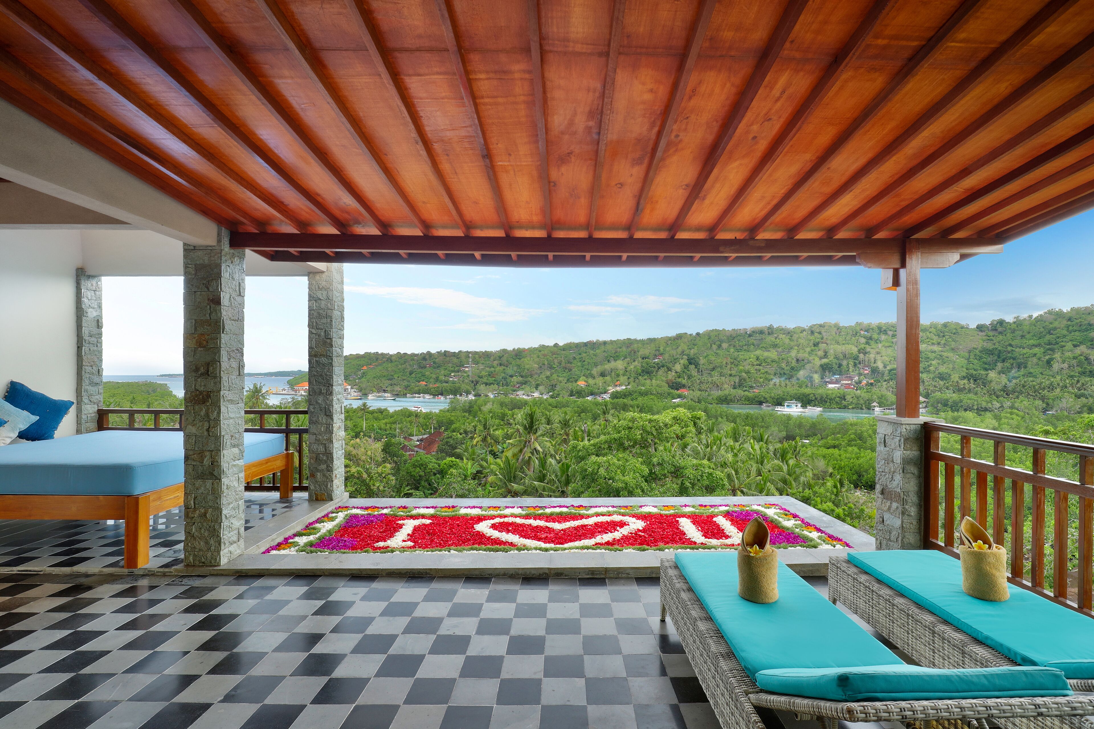 A balcony with a checkered floor, a bed with blue bedding, wicker furniture, and a view of a lush landscape.