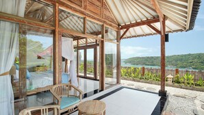 A room with a glass door leading to a balcony with a view of the sea and hills. There is a wooden chair and a small table on the balcony. - The Acala Shri Sedana (Lembongan Island)