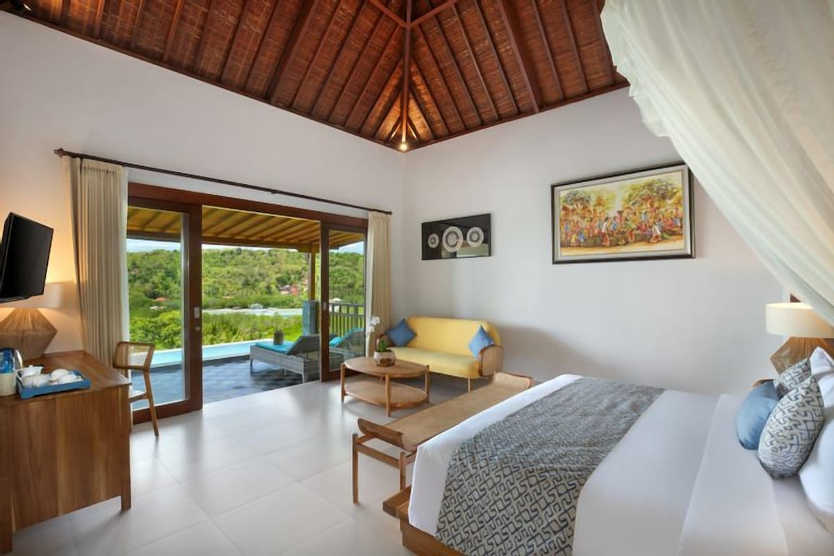 A bedroom with a bed, a sofa, a TV, and a desk. There is a balcony with a view of greenery.
