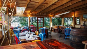 Een rustiek houten interieur met meerdere tafels en kleurrijke stoelen, grote ramen en een houten vat.