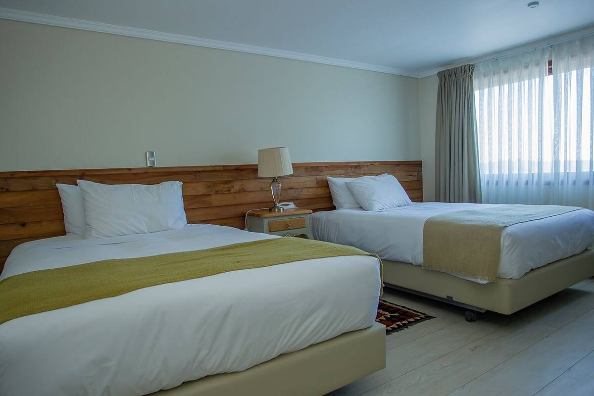Ein Hotelzimmer mit zwei Betten, Holz-Kopfteilen, weißer Bettwäsche, einem Nachttisch mit Lampe und einem Fenster mit Vorhängen.