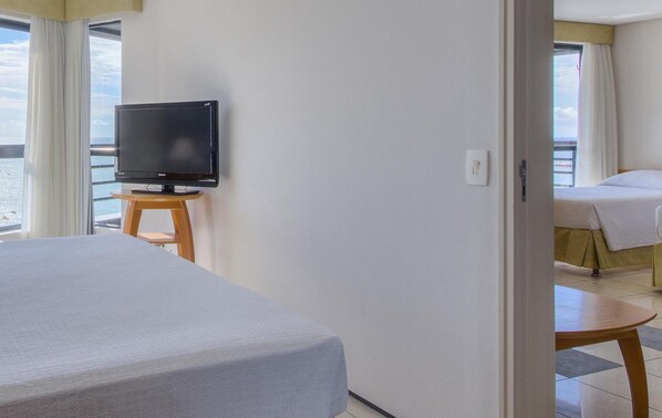 Una habitación de hotel con cama, televisión sobre una mesita y vista al mar a través de amplias ventanas.