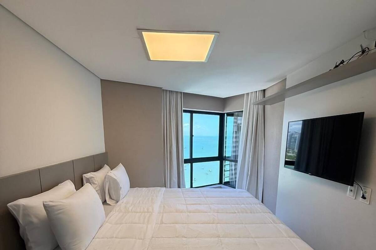 Ein großes Bett mit weißer Bettwäsche, eine Deckenleuchte, ein Fernseher an der Wand und Meerblick durch ein Fenster.