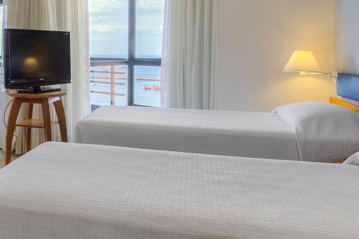 Ein Hotelzimmer mit zwei Betten, einem Fernseher auf einem Holztischchen, einer Lampe und Meerblick durch große Fenster.