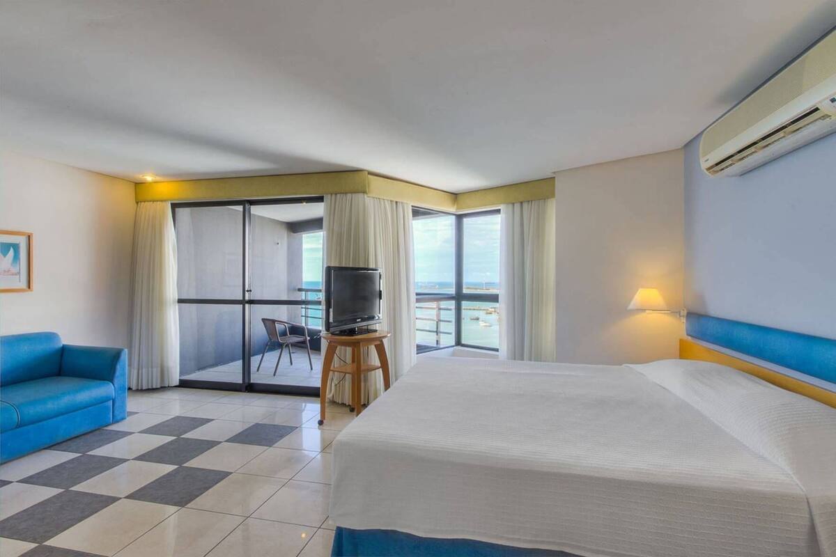 Ein Hotelzimmer mit Bett, Fernseher auf einem Holzgestell, blaues Sofa und Balkon mit Meerblick.