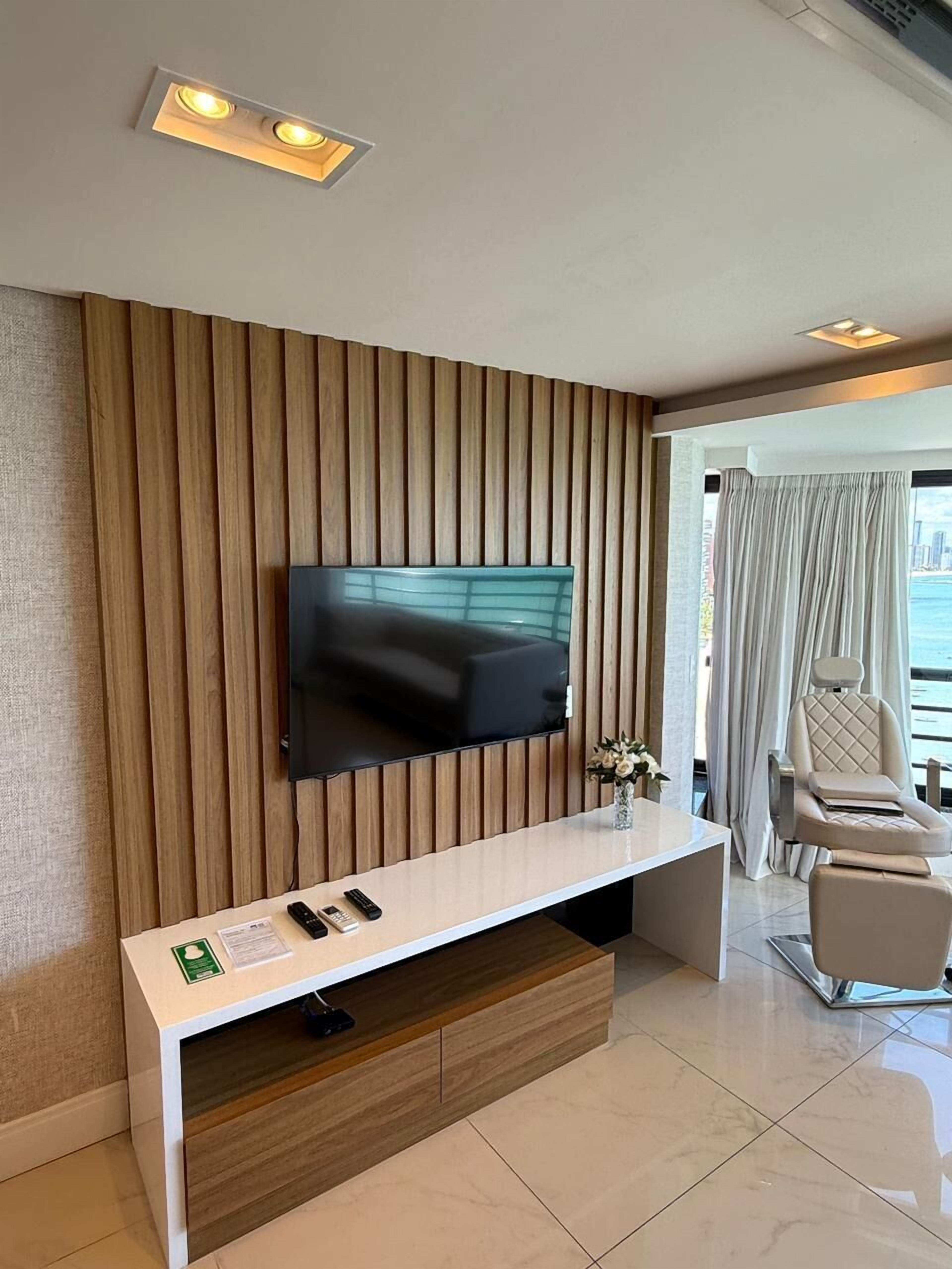 A modern living room with a flat-screen TV mounted on a wooden panel wall, a white console table with remote controls, and a balcony with a view.