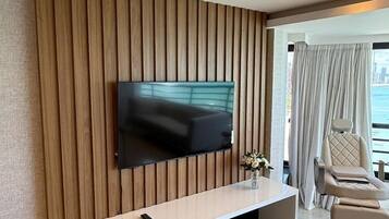 A modern living room with a flat-screen TV mounted on a wooden panel wall, a white console table with remote controls, and a balcony with a view.