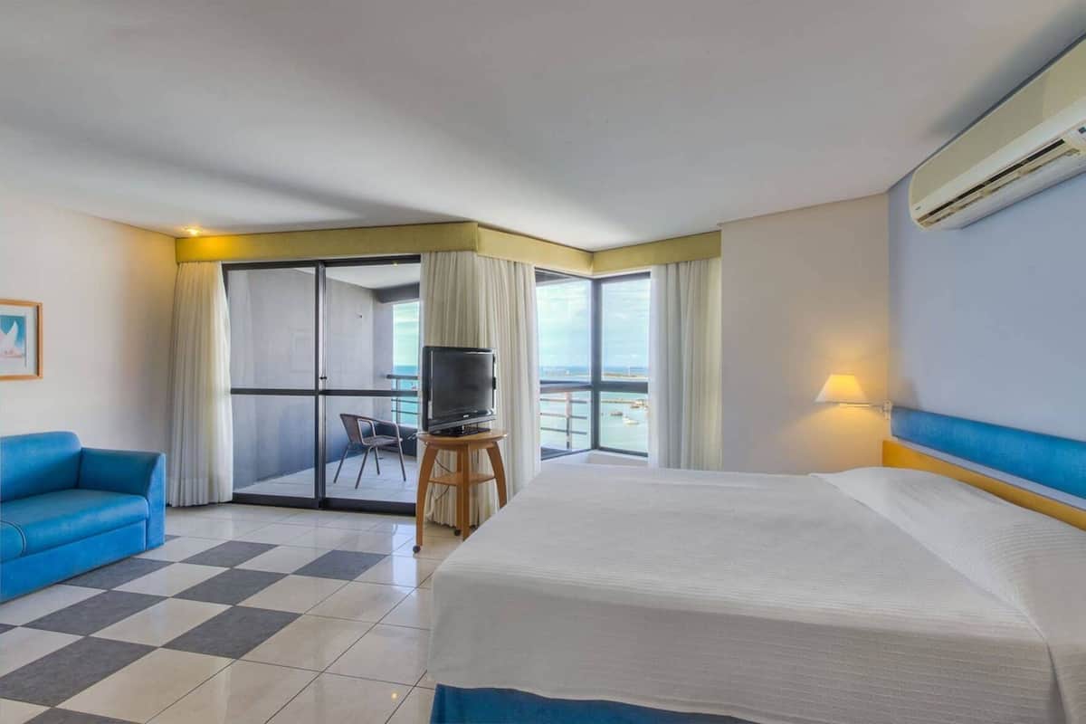 Ein Hotelzimmer mit Bett, Fernseher auf einem Holzgestell, blaues Sofa und Balkon mit Meerblick.
