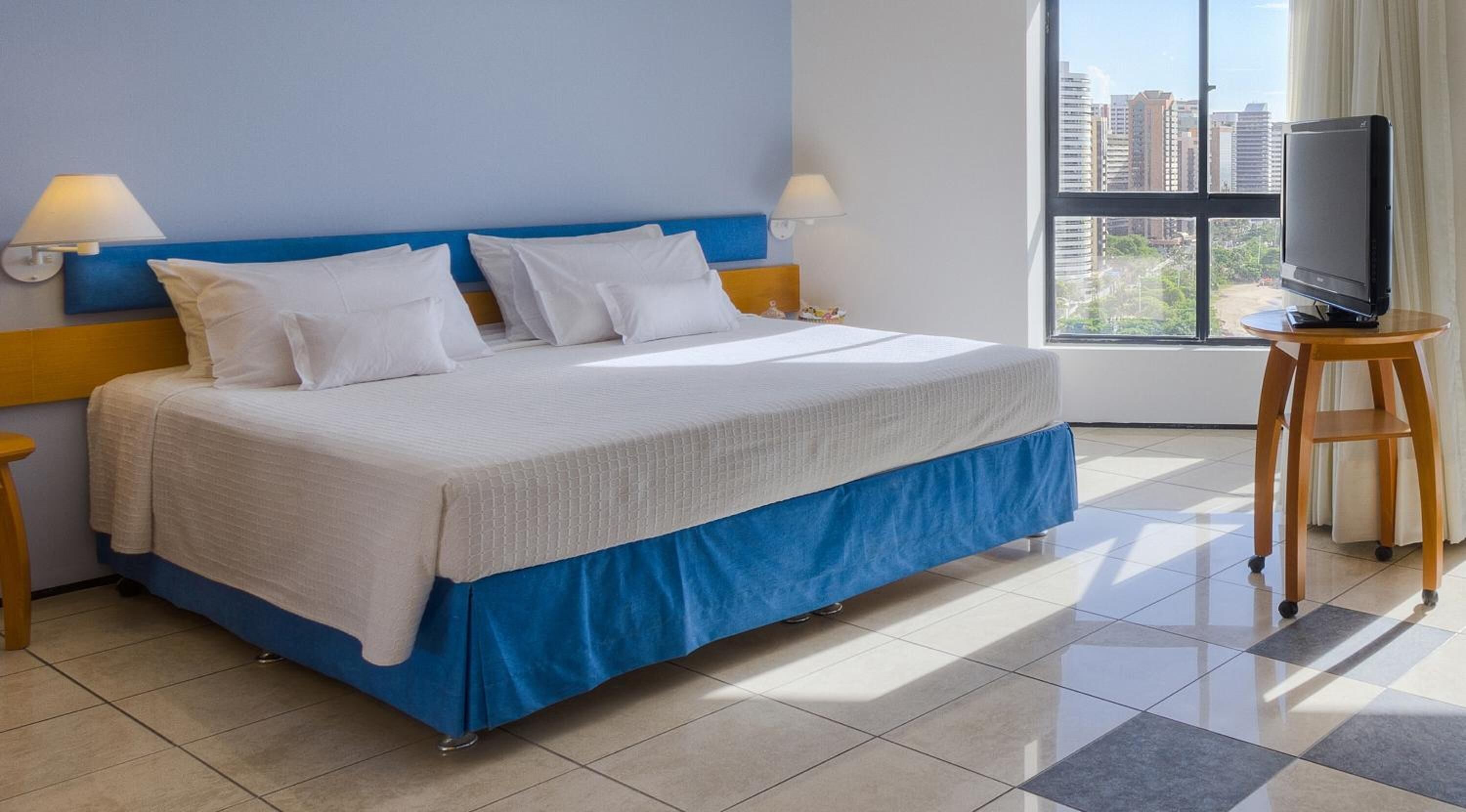 Ein Hotelzimmer mit Bett, zwei Nachttischlampen, einem Fernseher auf einem Holzboden und einem Fenster mit Blick auf die Stadt.