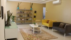 A living room with a yellow sofa, a dark sofa, a white coffee table, a glass side table, a bookshelf, and a large plant. - Family Suite DESA TEBRAU JOHOR BAHRU (Johor Bahru)