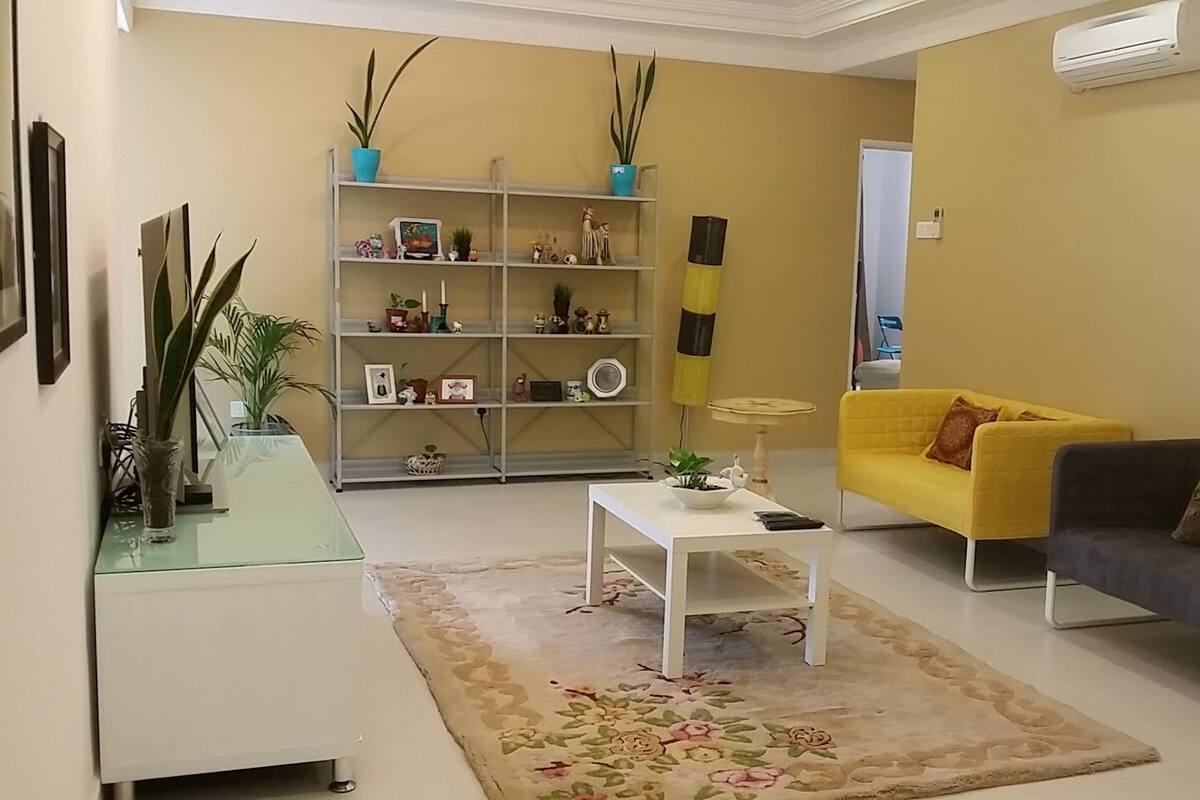 Ein Wohnzimmer mit einem gelben Sofa, einem dunklen Sofa, einem weißen Couchtisch, einem Glastisch, einem Bücherregal und einer großen Pflanze.
