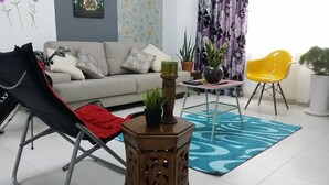 A living room with a sofa, a black folding chair, a small wooden table with a plant, a yellow chair, and a blue patterned rug. - Family Suite DESA TEBRAU JOHOR BAHRU (Johor Bahru)