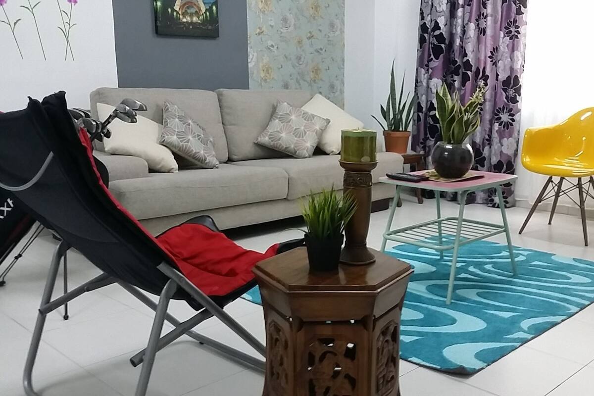 Ein Wohnzimmer mit einer Couch, einem schwarzen Klappstuhl, einem kleinen Holztisch mit einer Pflanze, einem gelben Sessel und einem blau gemusterten Teppich.