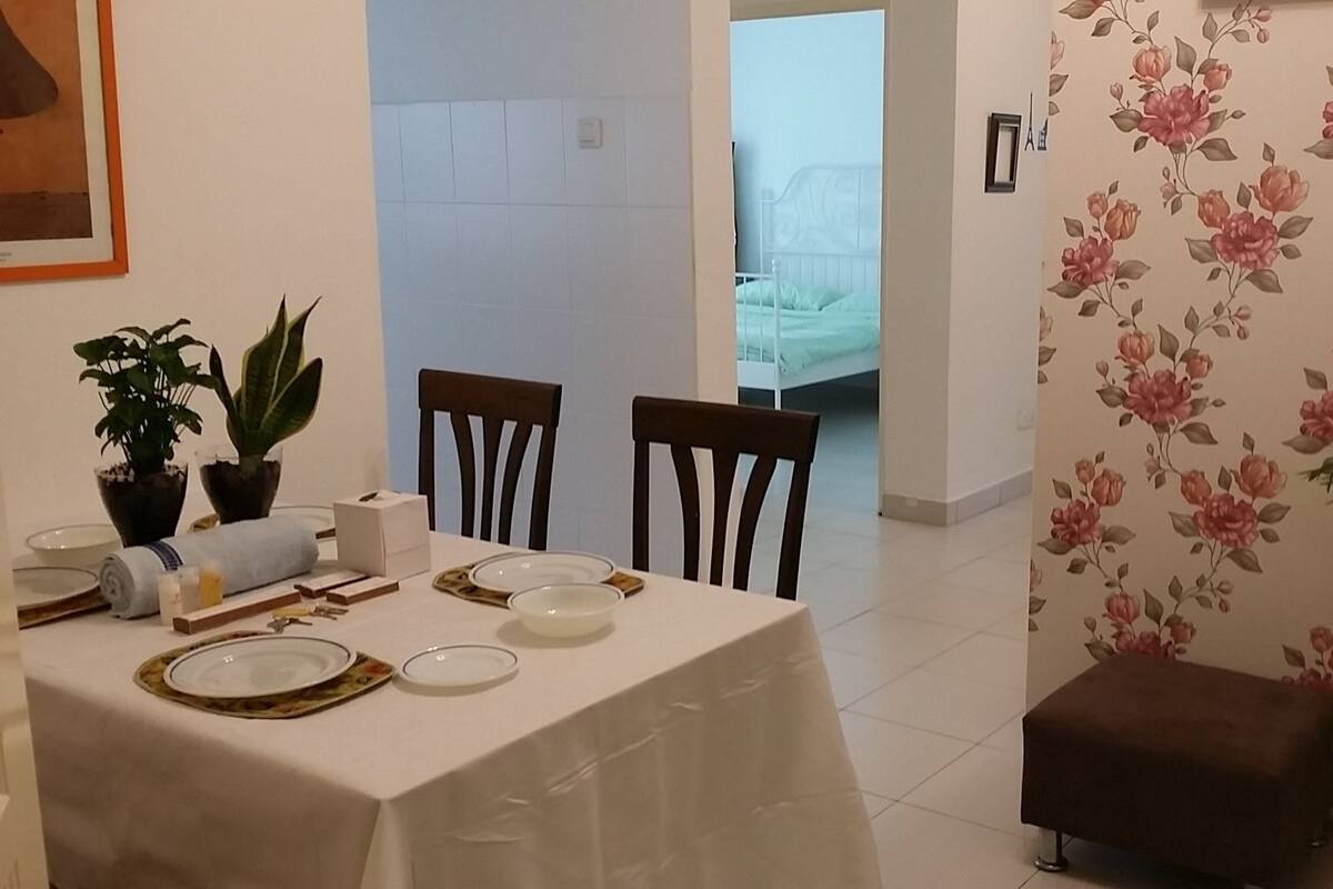 Ein Essbereich mit einem Tisch für zwei Personen, zwei Stühlen, einem kleinen braunen Hocker und zwei Blumentöpfen.