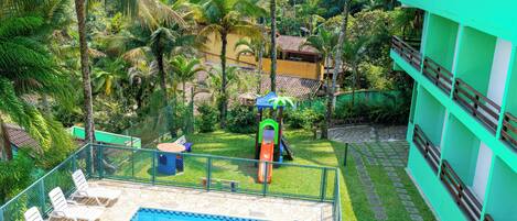 A swimming pool with lounge chairs, a children's play area, and a green building with a balcony.