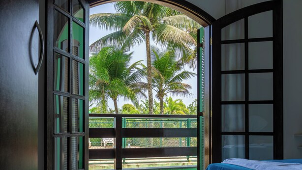 Una habitación con una cama, un ventanal con vista a palmeras y un balcón con barandilla verde.