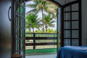 Quarto com cama, janela vista para palmeiras e varanda com grade verde.