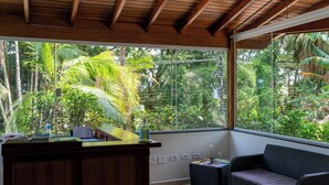 Una habitación con un escritorio de madera, un sofá de mimbre y un amplio ventanal con vistas a un follaje exuberante.