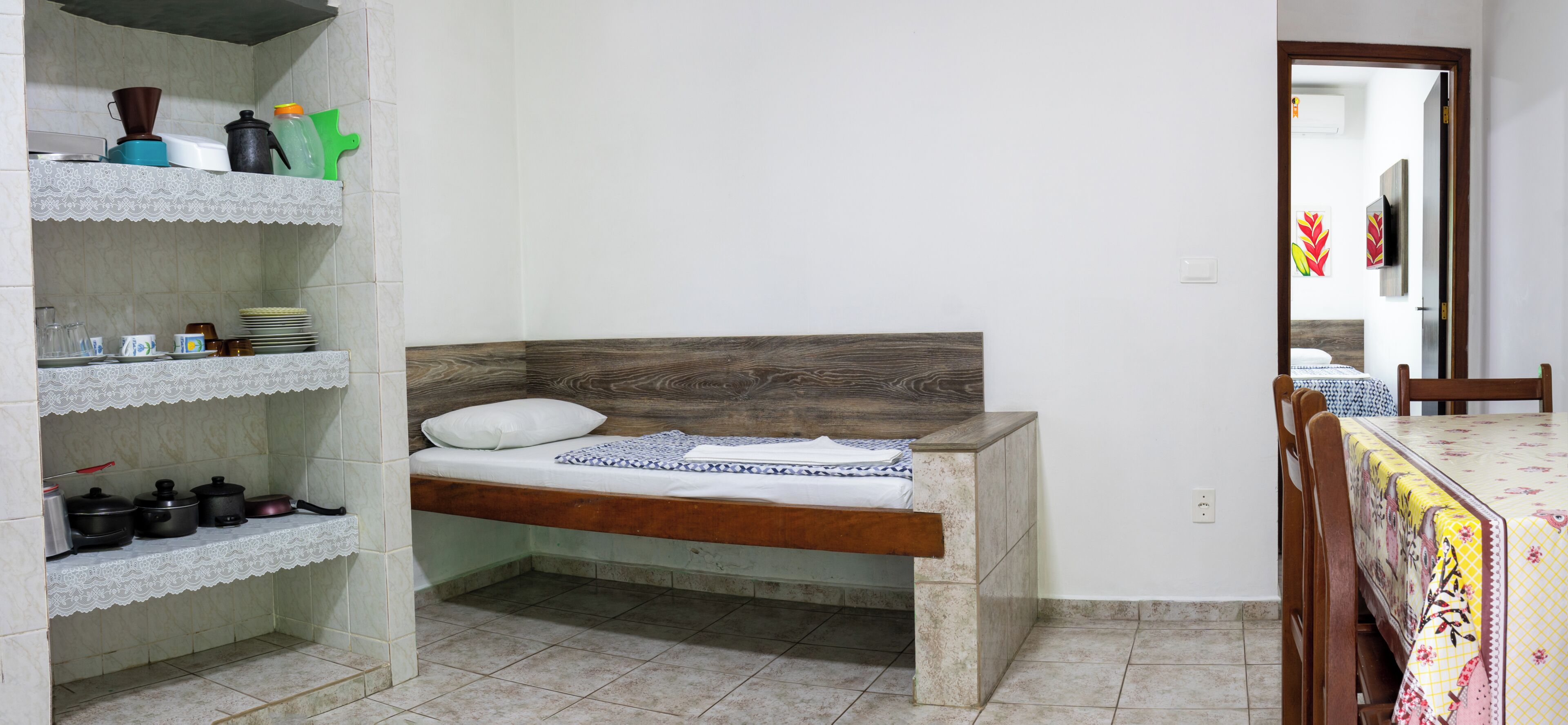 A bedroom with a bed, a shelf with kitchen items, and a dining table with chairs.
