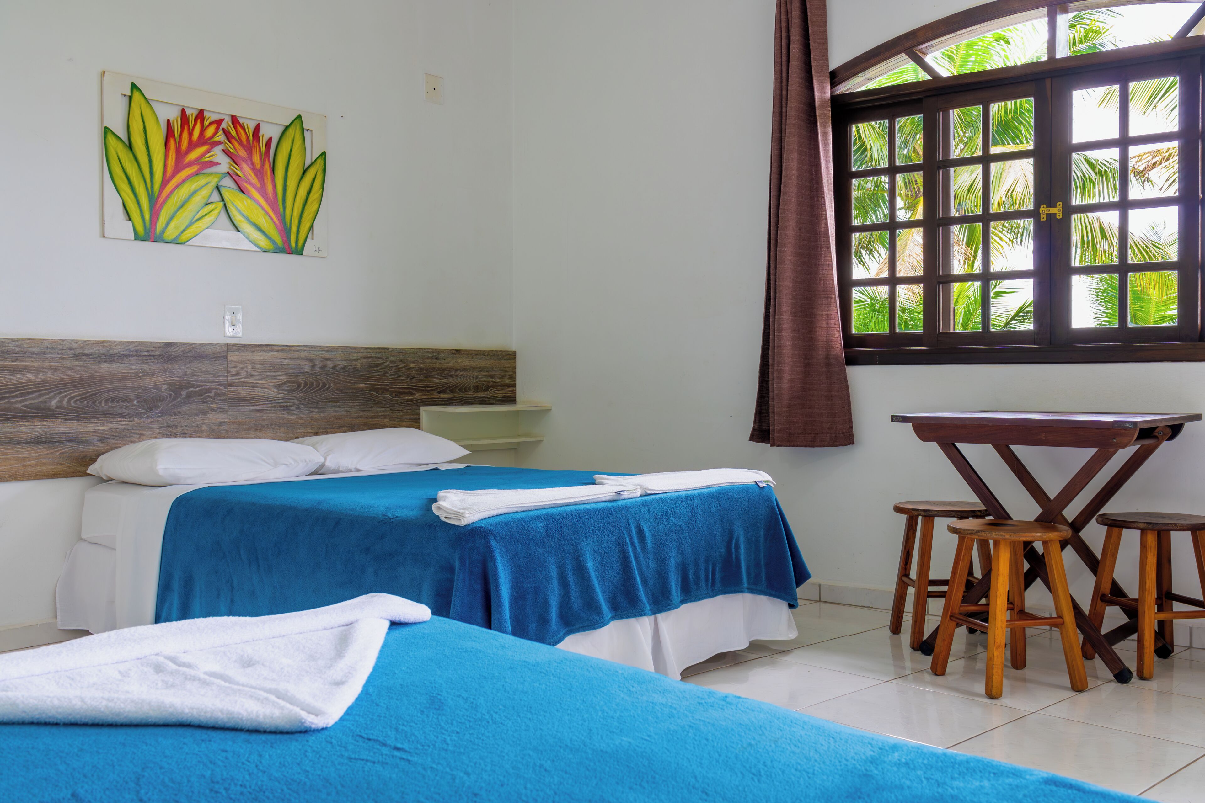 Una habitación con dos camas, un cabecero de madera, una ventana con vista a palmeras y una pequeña mesa de madera con taburetes.