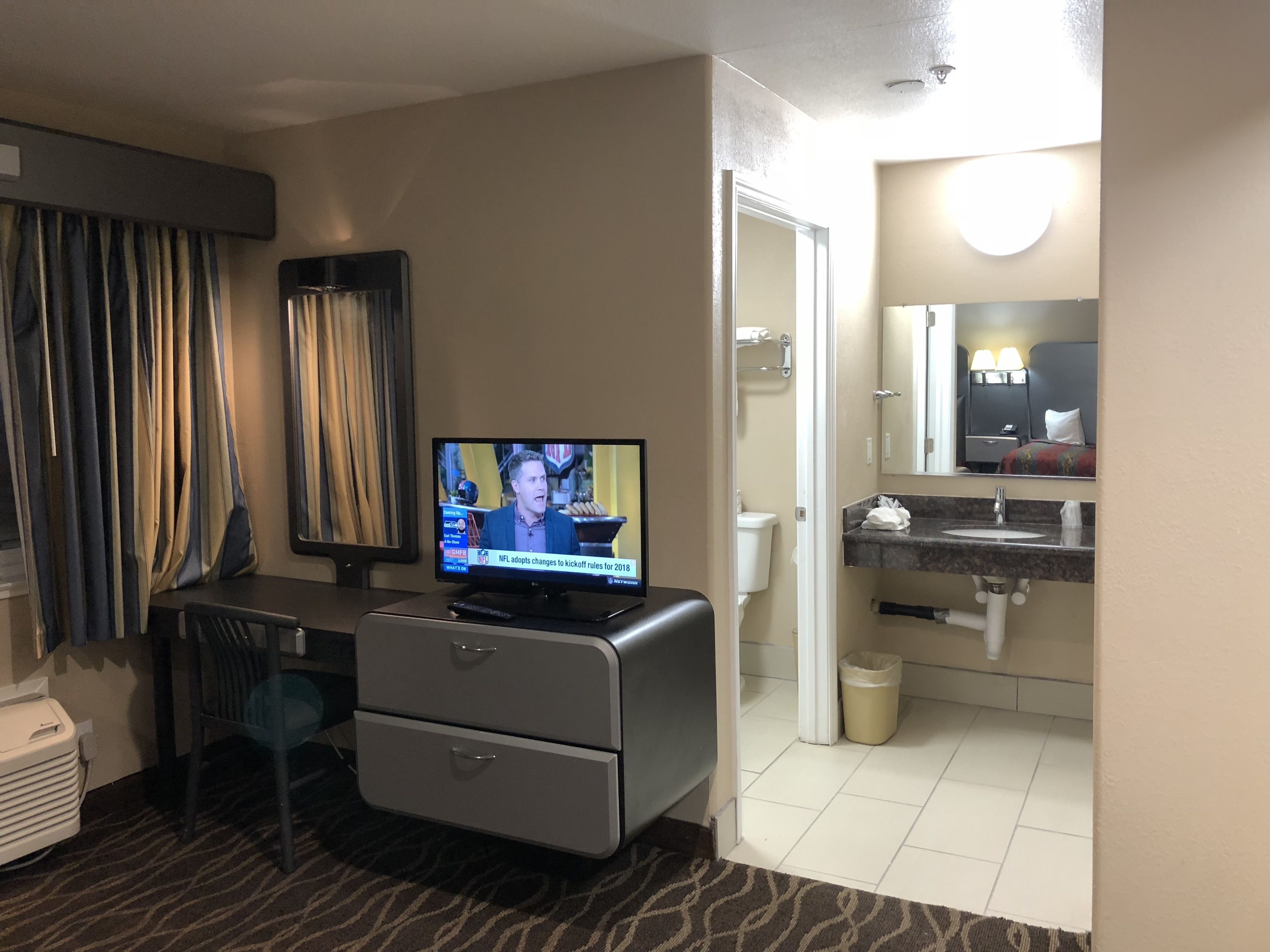 Une chambre d’hôtel équipée d’une télévision diffusant un programme d’information, d’un bureau avec une chaise, d’une commode, d’une salle de bain avec un lavabo et un miroir, et d’une poubelle.