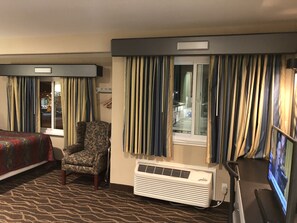 A hotel room with a patterned carpet, a chair, an air conditioning unit, and a television.