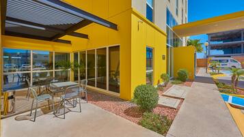 A modern building with yellow exterior walls, large windows, and outdoor seating area with metal chairs and tables.