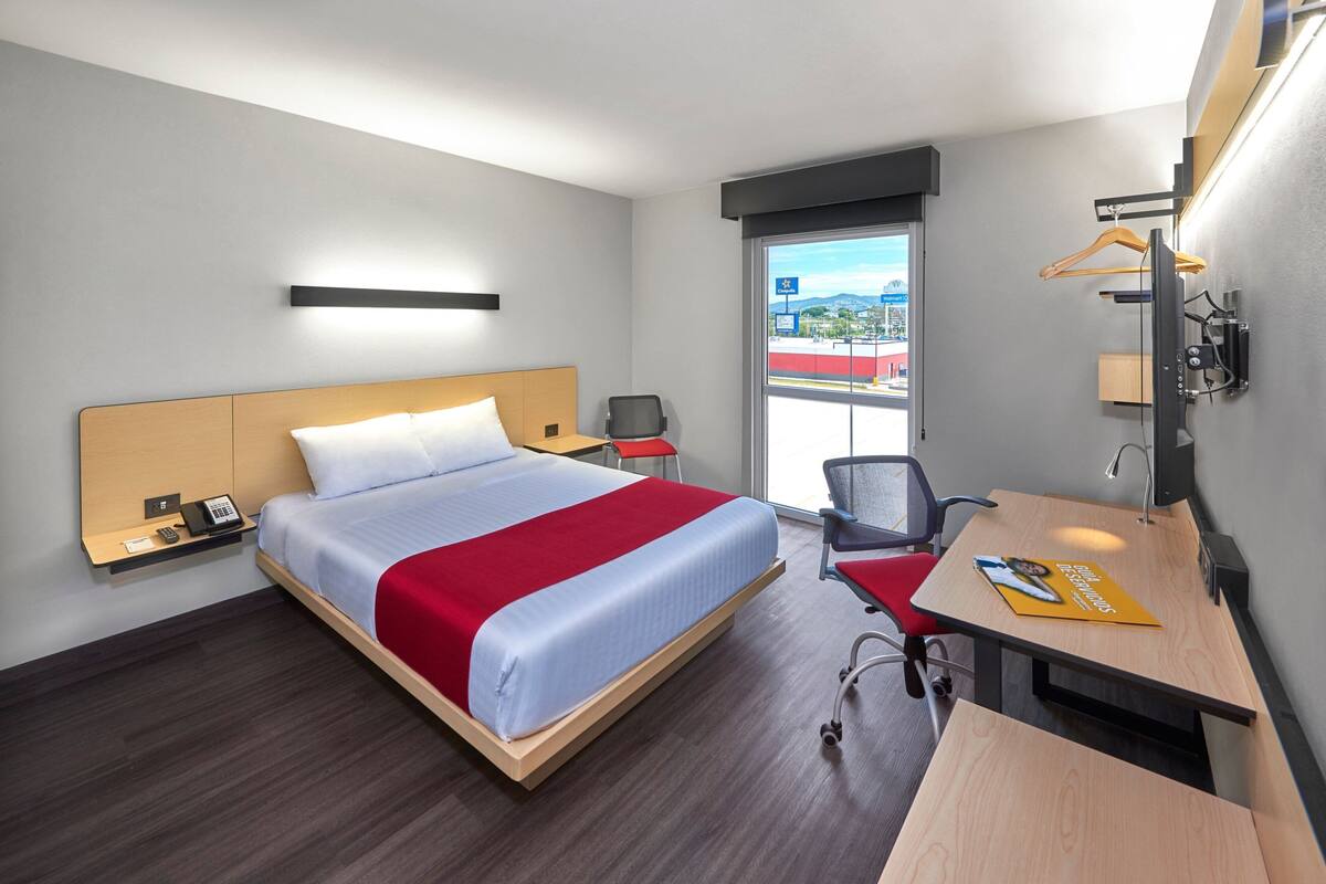Habitación de hotel con cama, escritorio, silla, teléfono y vistas al exterior.