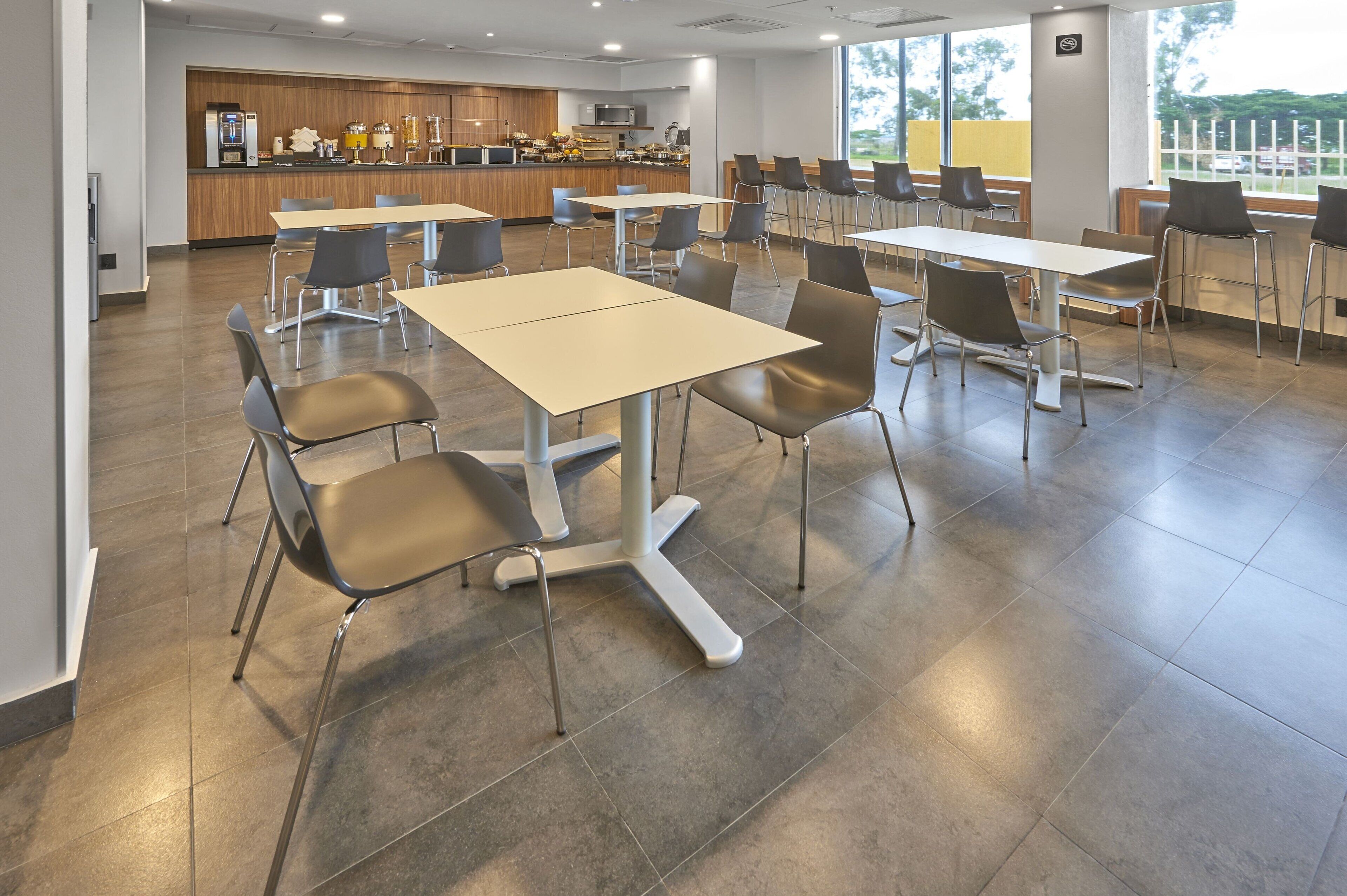 A modern dining area with multiple tables and chairs, a kitchen in the background, and large windows providing natural light.
