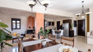 A spacious living area with a dining table set for four, a fireplace, and a kitchen in the background. - Villa Fanouris (Chania)