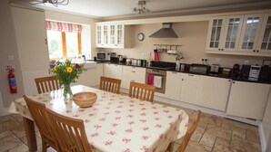 Une cuisine avec des meubles blancs, une table à manger recouverte d’une nappe à motifs floraux, des chaises en bois et un vase rempli de fleurs.