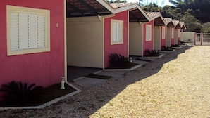 Una hilera de edificios pequeños de un piso, con un estilo arquitectónico uniforme, que presenta fachada roja y adornos color beige. Cada edificio tiene una pequeña entrada con puerta y una ventana con persianas. El suelo está cubierto de grava, y hay plantas a los pies de los edificios.