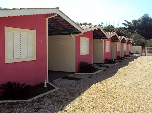 Une rangée de petits bâtiments d’un seul étage, de style architectural uniforme, se distingue par sa façade rouge et ses ornements beiges. Chaque bâtiment possède une petite entrée avec une porte et une fenêtre dotée de volets. Le sol est recouvert de gravier et des plantes sont disposées au pied des bâtiments.