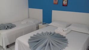 Habitación de hotel con dos camas individuales, cada una con cubrecama y almohadas blancas. Hay una mesita de noche con un control remoto y una toalla doblada. La habitación tiene una pared de acento azul y un techo blanco.