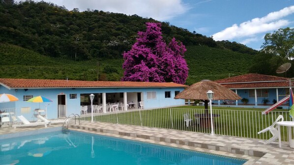 A swimming pool area with lounge chairs, a thatched-roof hut, and a building with a red roof. - Pousada Primavera da Serra (Serra Negra)