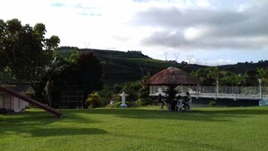 A grassy area with a thatched-roof gazebo, a white statue, and a bridge.