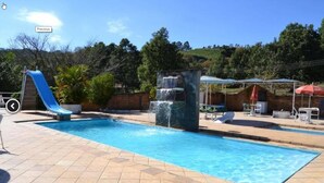 A swimming pool with a slide and a waterfall feature, surrounded by a tiled deck and outdoor furniture. - Pousada Sitio Bom Fim (Serra Negra)