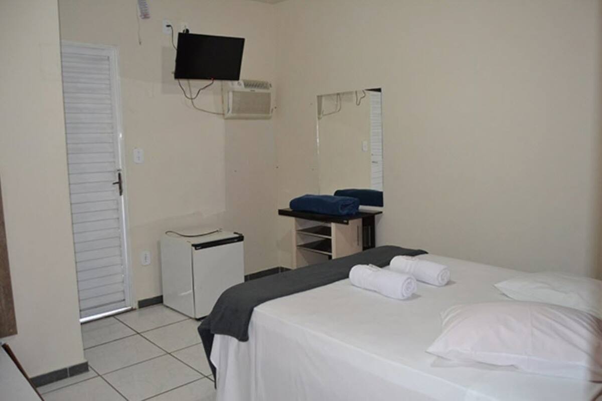 Quarto de hotel com uma cama, uma pequena escrivaninha, uma cadeira, televisão fixada na parede e um espelho.
