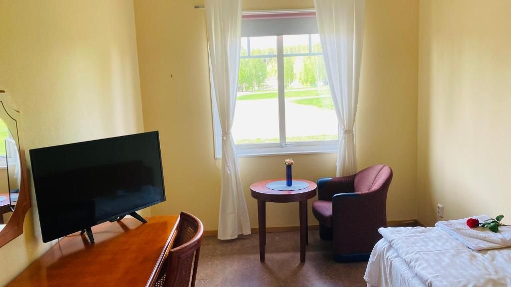 A room with a television on a wooden stand, a small round table with a blue object, a pink chair, and a bed with a white bedspread and a red rose.