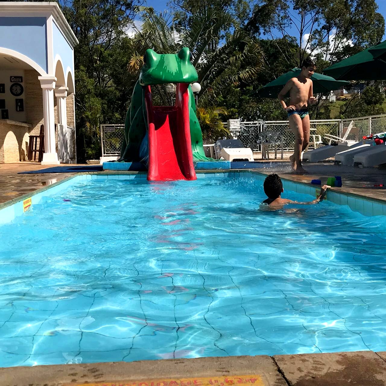 Una piscina con tobogán, rodeada por una zona vallada con sillas de descanso y sombrillas.