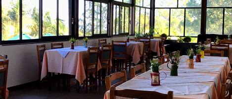 A dining area with tables set for guests, large windows with a view of palm trees, and wooden chairs.