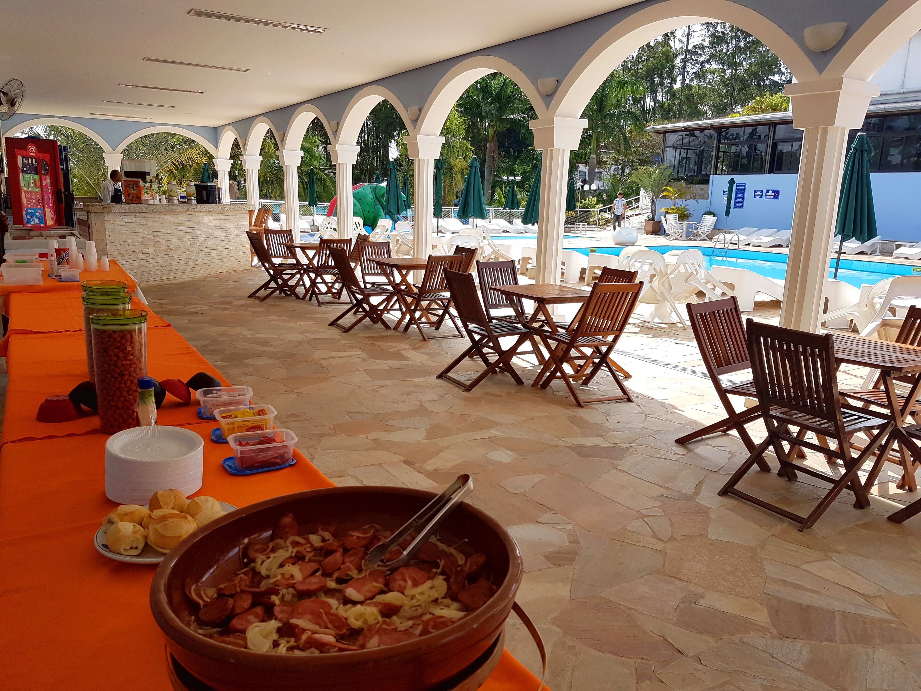 Ein überdachter Essbereich im Freien mit Tischen und Stühlen, ein Büfett mit Speisen sowie ein Pool im Hintergrund.