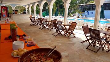 A covered outdoor dining area with tables and chairs, a buffet table with food items, and a pool in the background.
