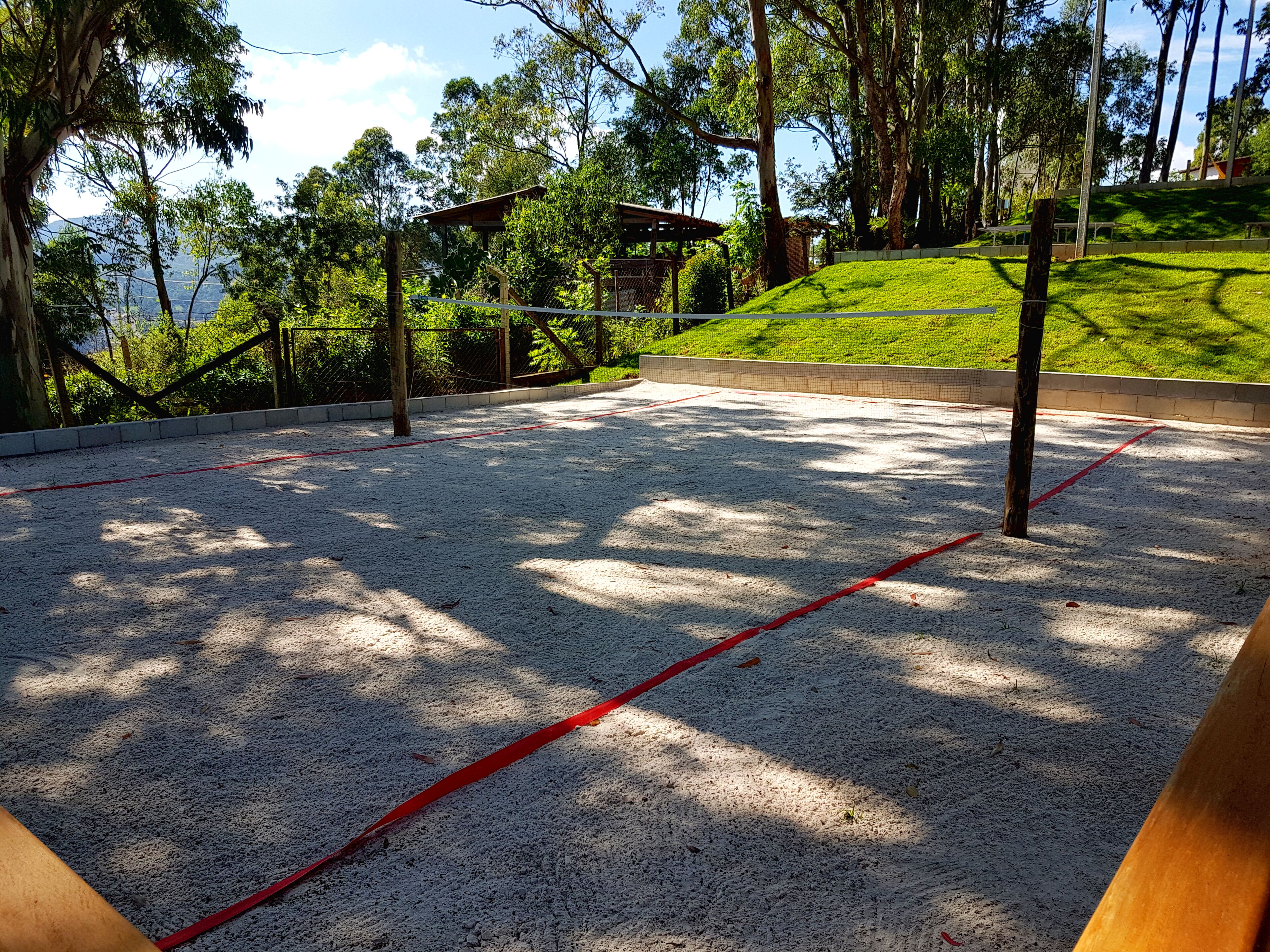 Una cancha de voleibol con una línea divisoria roja, rodeada de árboles y una pendiente cubierta de césped.