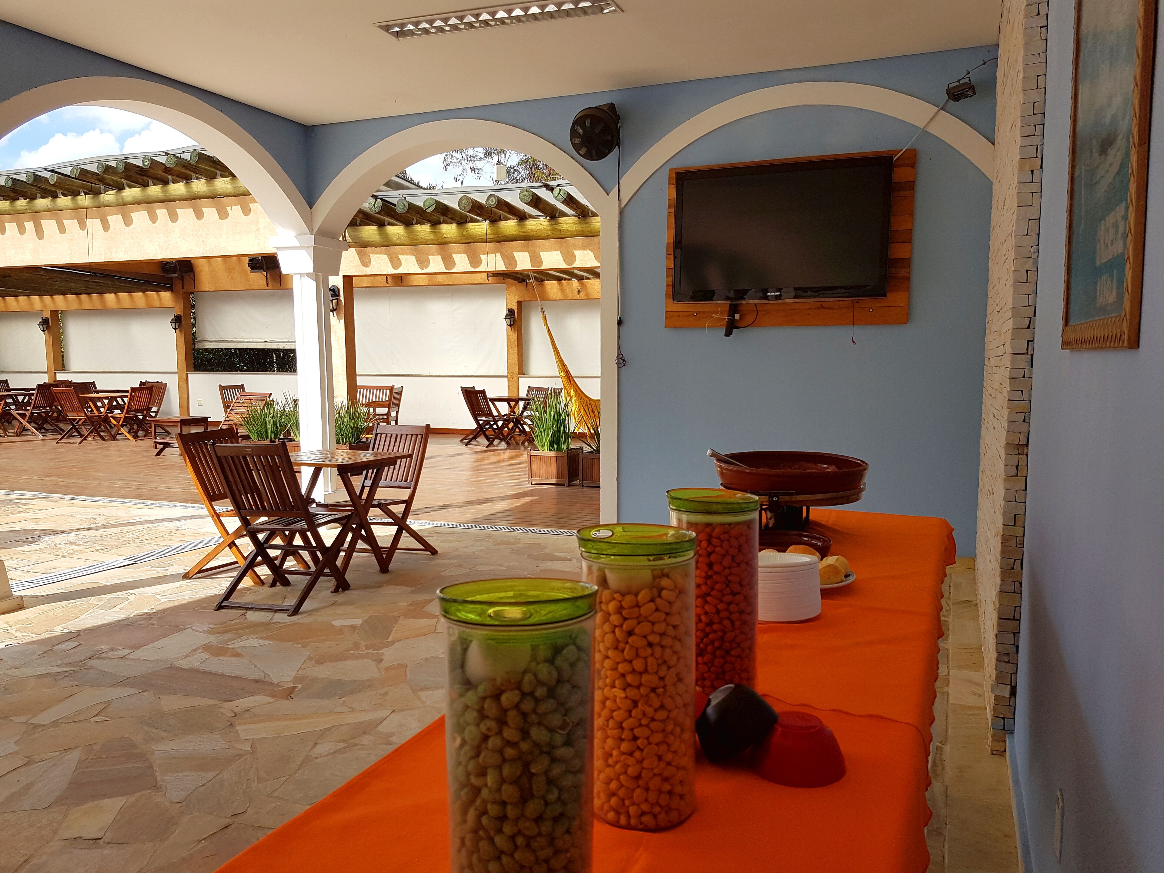 Uma área de estar externa com mesas e cadeiras, uma TV fixada na parede e potes de lanches sobre uma mesa.