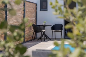 A small outdoor seating area with two chairs and a table, a potted plant, and a pool in the background. - John Mary Apartments (Hersonissos)