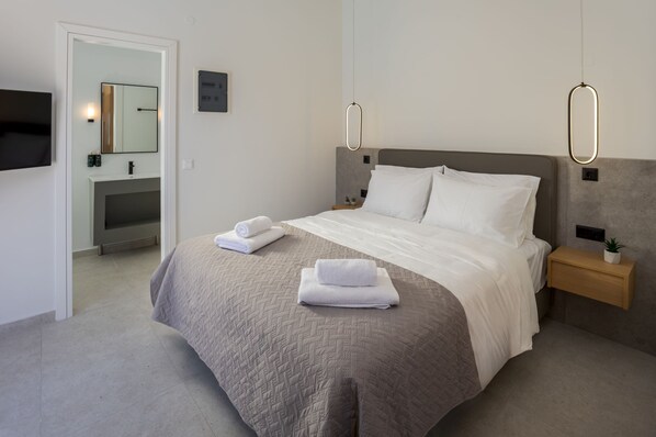 A modern hotel room with a large bed, a bedside table with a plant, and a built-in TV. - John Mary Apartments (Hersonissos)